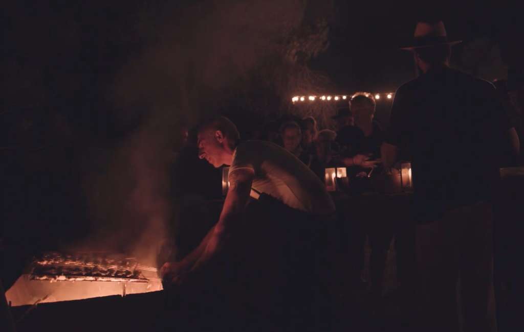 Man leaning over a glowing barbecue grill at night, with onlookers and string lights behind