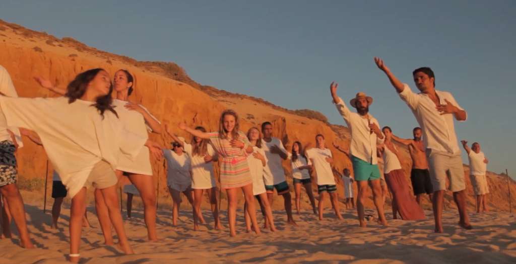 Group in white clothes stretching their arms on a beach below orange cliffs at sunset