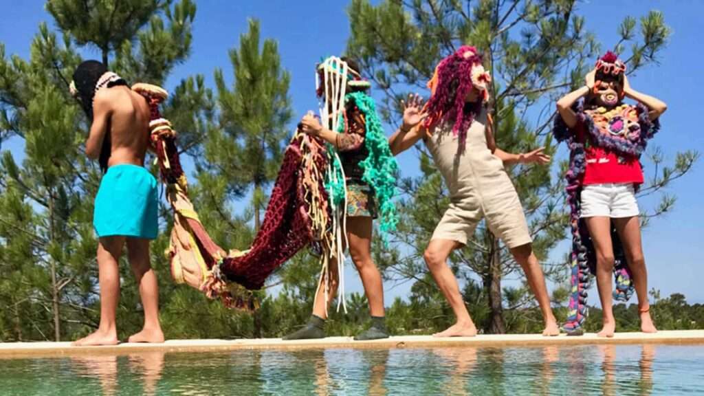 Four people in colorful knitted masks and costumes posing on a pool edge with trees behind