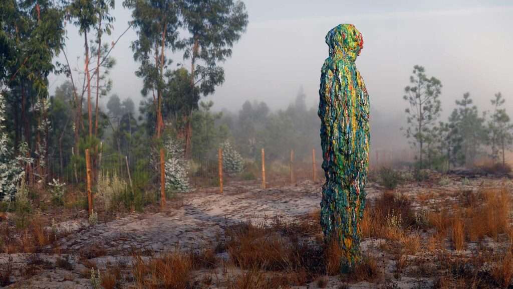 Tall sculpture covered in multicolored paint drips standing in misty pine woodland