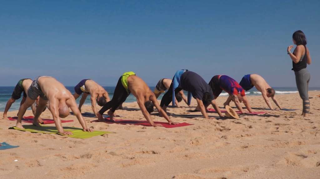 Group in downward dog yoga pose on beach mats while an instructor watches