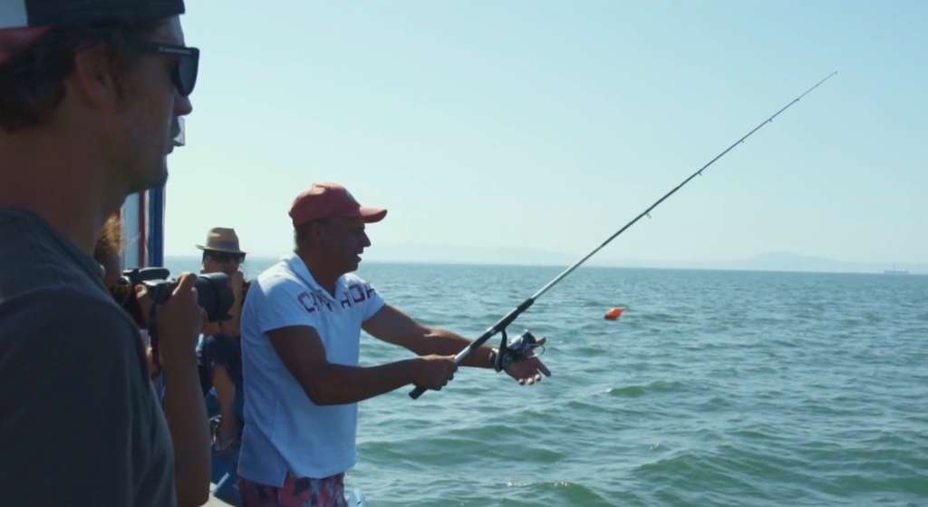 Man in a red cap fishing from a boat while another person films with a camera