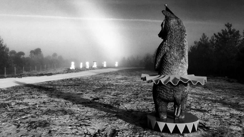 Black and white photo of a bear sculpture in a tutu on a misty path with glowing figures