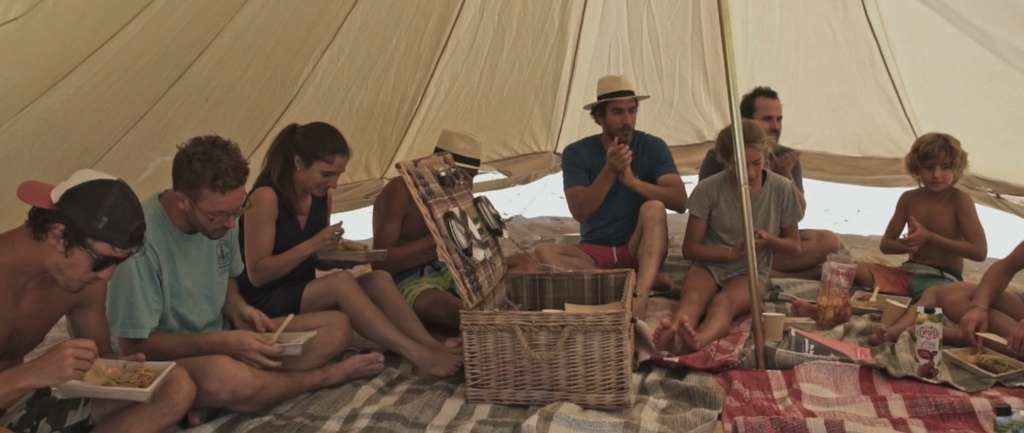 People sitting on blankets eating inside a canvas tent, with a wicker picnic basket open