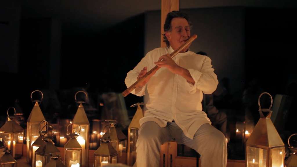 Man in a white shirt playing a bamboo flute surrounded by candle lanterns