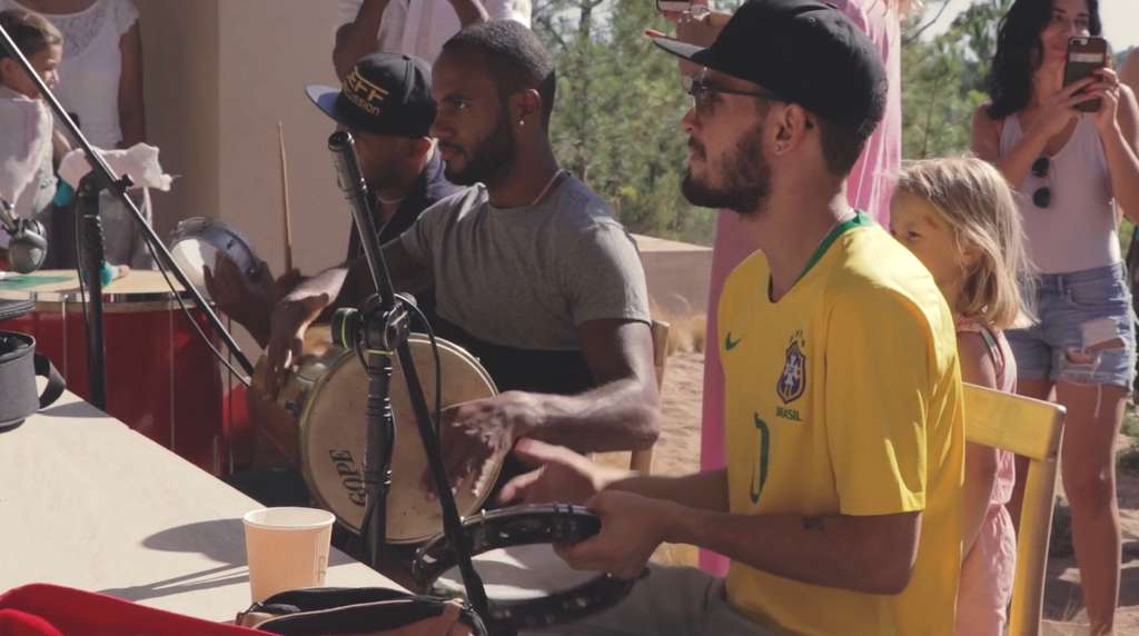 Musicians playing drums and a tambourine outdoors, with children and onlookers behind