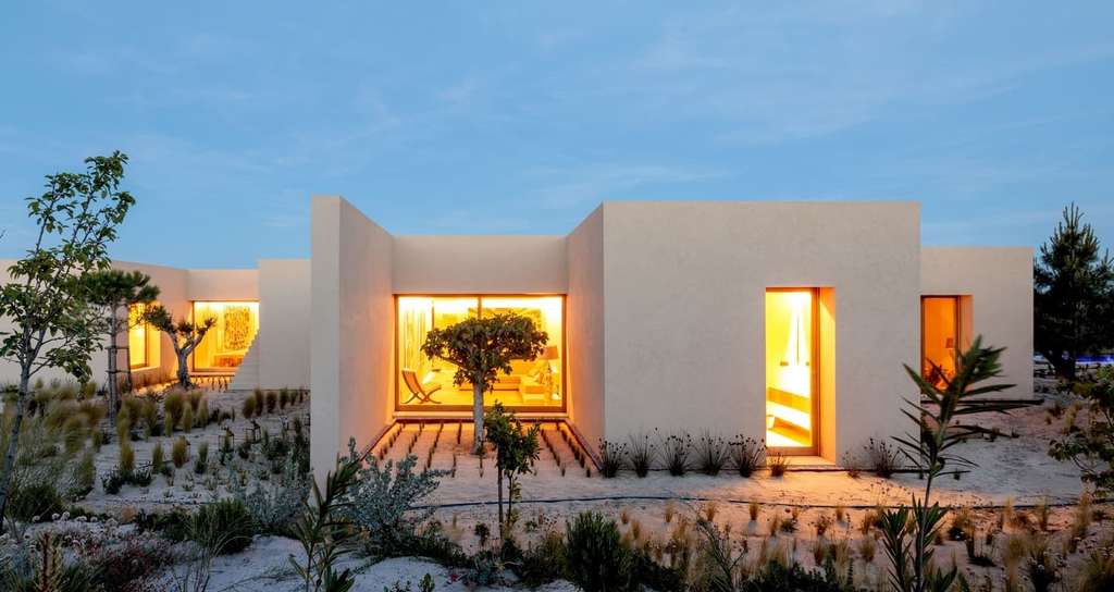 Sand-colored single-storey house glowing with warm light at dusk, sandy garden in front