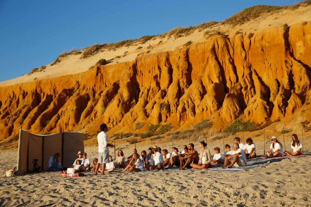 People in white seated on the beach in front of tall orange cliffs, one person standing