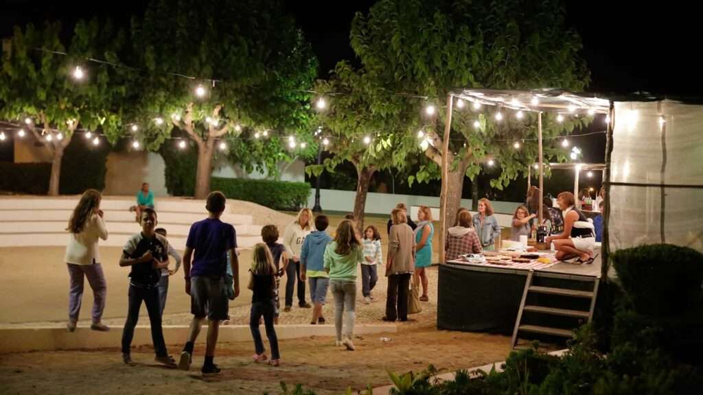 Families and children gathered in an outdoor square at night under string lights, with a food table