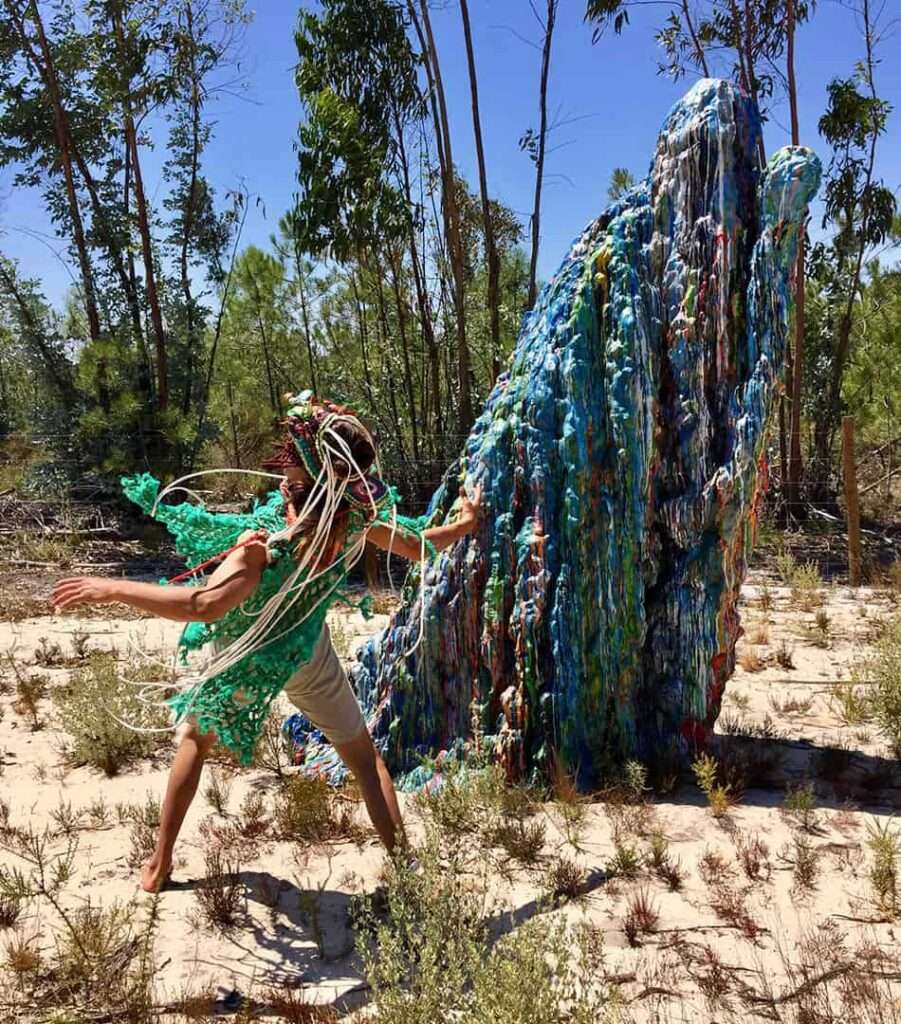 Person in green knitted costume and headpiece beside a large blue and orange fibre sculpture