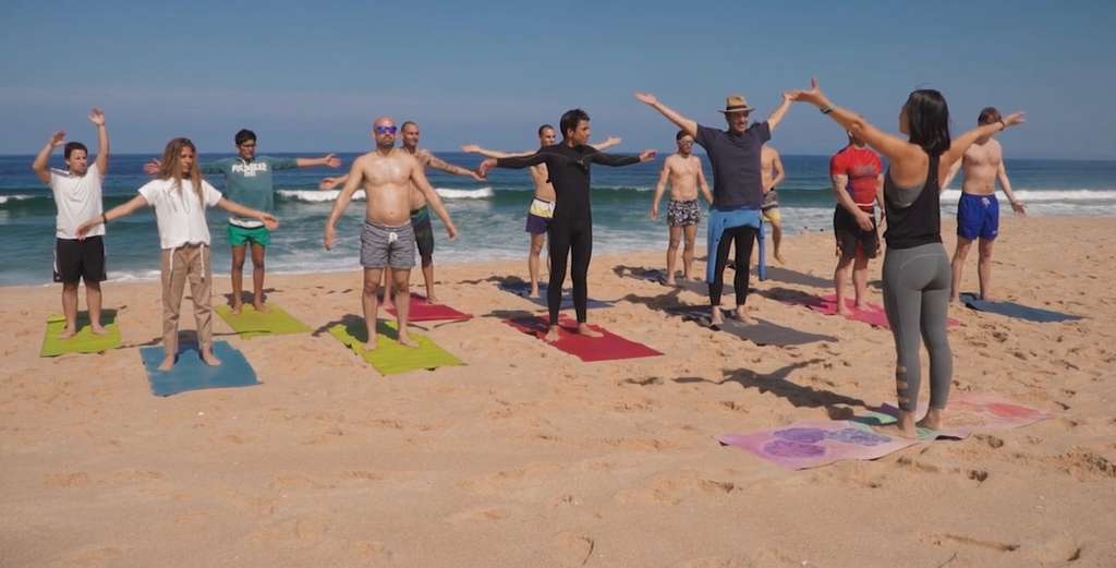 Group standing with arms outstretched on colorful mats on a beach, led by an instructor