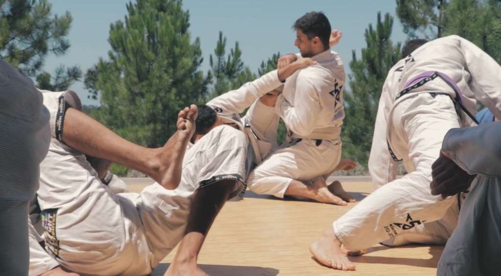 Group in white jiu-jitsu uniforms practicing grappling on a mat outdoors among pine trees