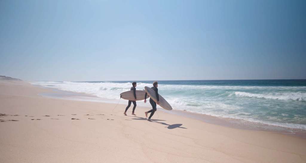 Two people in wetsuits carrying surfboards along a sandy beach toward the waves